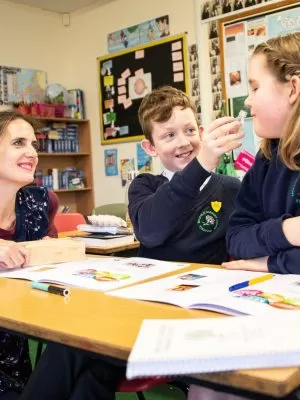 Two school children sitting at a desk doing a mindfulness activity, with a teacher crouched down beside their desk.