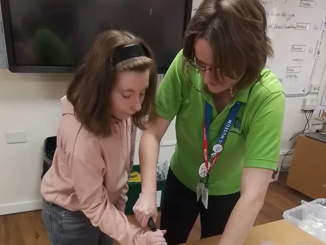 An adult standing beside a young person, helping them with a craft activity on a table.