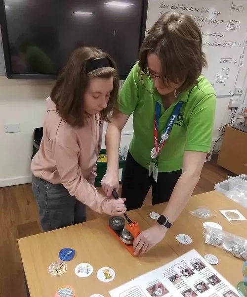 An adult standing beside a young person, helping them with a craft activity on a table.