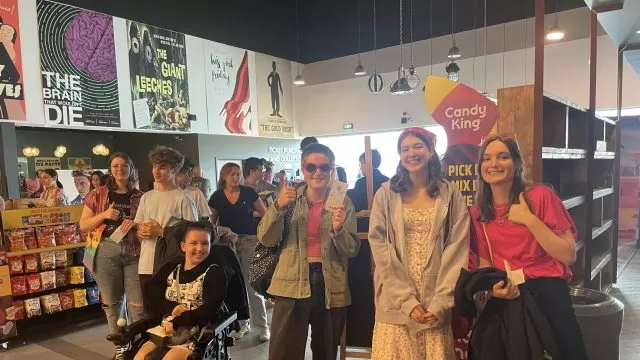 Five young people standing and one young person in a wheelchair, in a cinema, facing the camera and smiling.