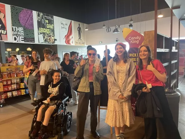 Five young people standing and one young person in a wheelchair, in a cinema, facing the camera and smiling.