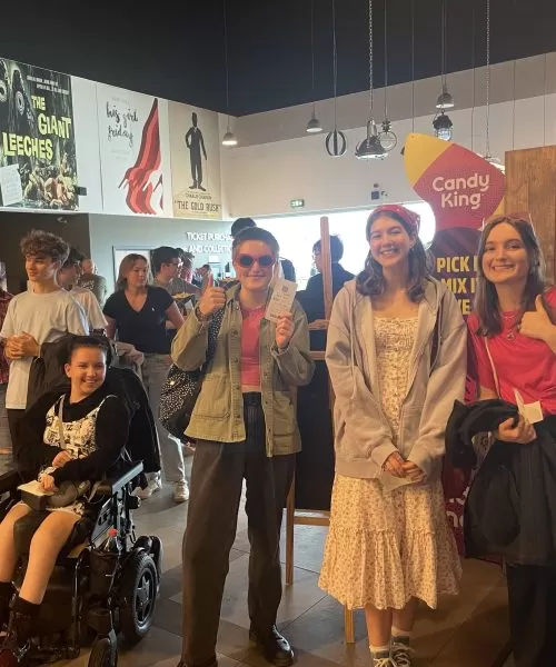 Five young people standing and one young person in a wheelchair, in a cinema, facing the camera and smiling.