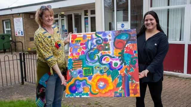 Two people standing holding a colourful painting between them.
