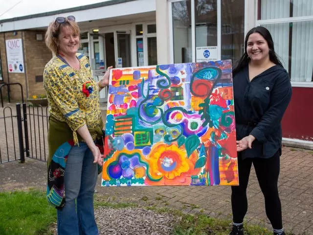 Two people standing holding a colourful painting between them.