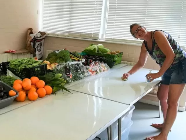 A woman smiling and standing with a cleaning cloth and sanitiser in front of a white table with fruit and vegetables.