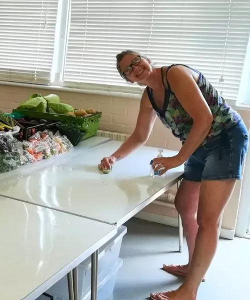 A woman smiling and standing with a cleaning cloth and sanitiser in front of a white table with fruit and vegetables.