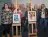 People gathered around two pieces of art on easels. They are all facing the camera.