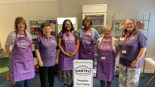 6 ladies in purple tops or aprons standing around a white food pantry sign.