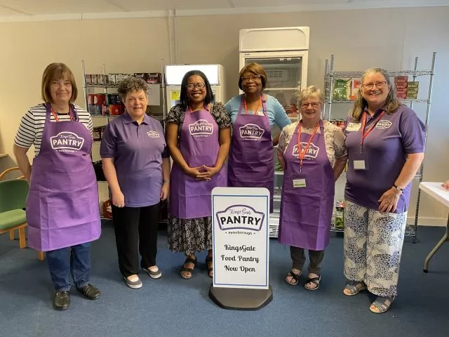 6 ladies in purple tops or aprons standing around a white food pantry sign.