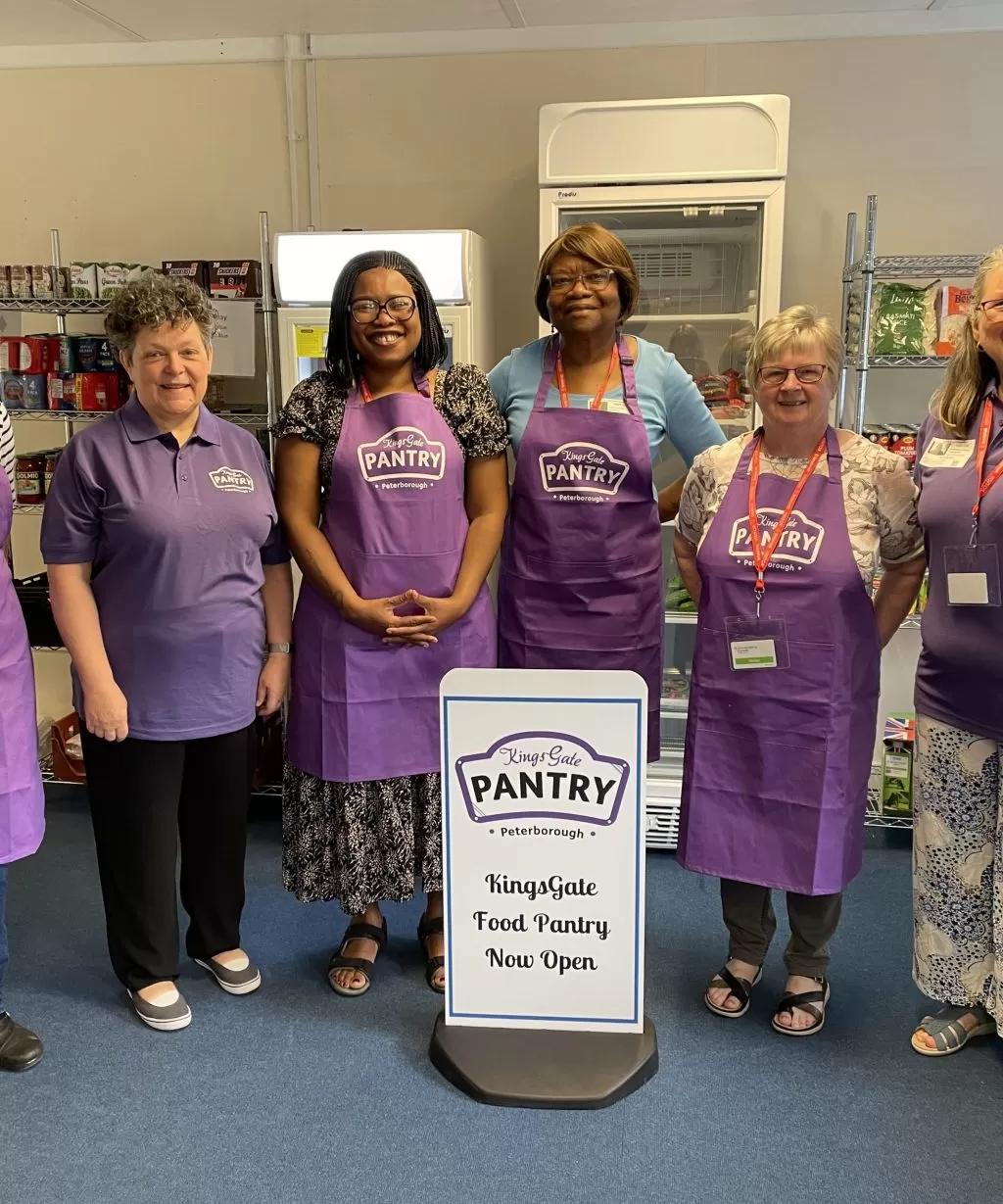 6 ladies in purple tops or aprons standing around a white food pantry sign.