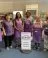 6 ladies in purple tops or aprons standing around a white food pantry sign.