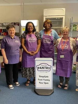 6 ladies in purple tops or aprons standing around a white food pantry sign.