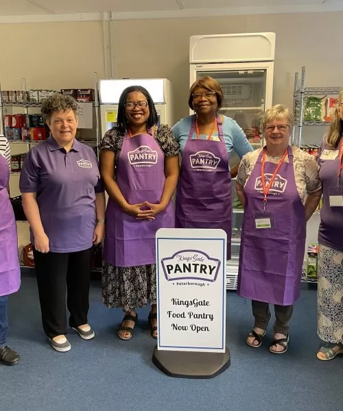 6 ladies in purple tops or aprons standing around a white food pantry sign.