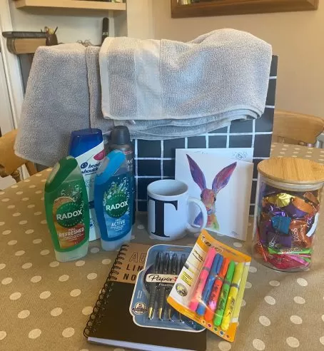 A welcome pack on top of a table, including towels, toiletry, a mug, a card, sweets in a jar, pens and a notepad.