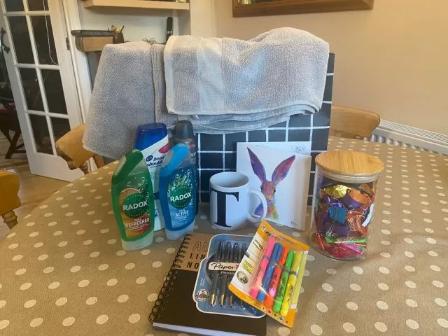 A welcome pack on top of a table, including towels, toiletry, a mug, a card, sweets in a jar, pens and a notepad.