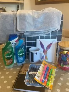 A welcome pack on top of a table, including towels, toiletry, a mug, a card, sweets in a jar, pens and a notepad.