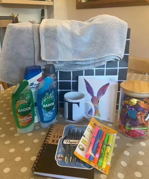 A welcome pack on top of a table, including towels, toiletry, a mug, a card, sweets in a jar, pens and a notepad.