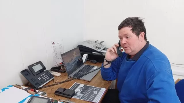 A person in a blue jumper answering a phone at a desk.