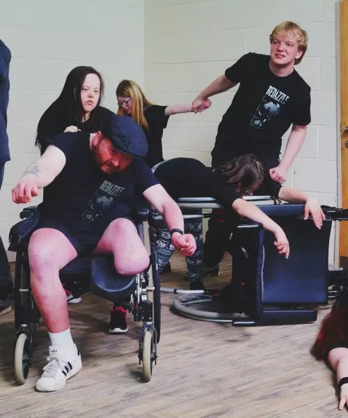 Actors mid-action. Two are holding hands. One is lying on the floor. One person is on a wheelchair, with another wheelchair pushed over with the person leaning on it.