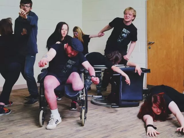 Actors mid-action. Two are holding hands. One is lying on the floor. One person is on a wheelchair, with another wheelchair pushed over with the person leaning on it.