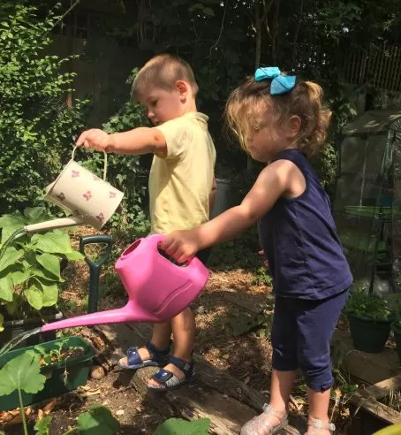 Two children watering plants in a garden with watering cans.