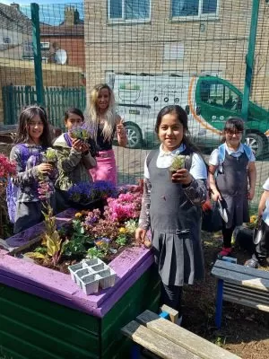 A group of children and an adult stood around a raised flower bed holding plants.