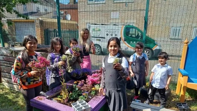 A group of children and an adult stood around a raised flower bed holding plants.