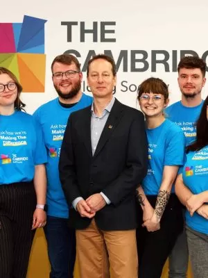 A man in a suit surrounded by people in blue shirts with The Cambridge Building Society's logo. They are standing in front of a white wall with The Cambridge's logo.