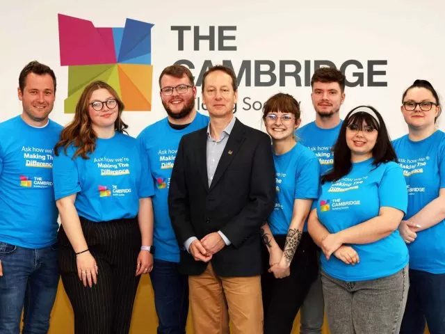 A man in a suit surrounded by people in blue shirts with The Cambridge Building Society's logo. They are standing in front of a white wall with The Cambridge's logo.