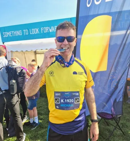 A runner standing in front of a flag with the charity's logo, wearing a yellow, branded running top with a running number. They are looking at the camera, biting their medal.